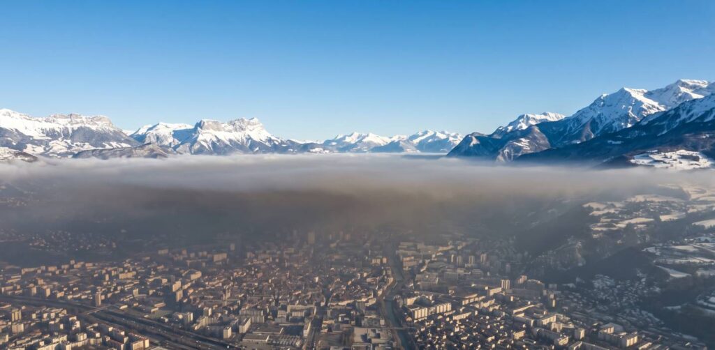 Photo aérienne de l'agglomération grenobloise en situation d'inversion thermique hivernale, nappe de pollution visible flottant au-dessus de la ville, montagnes enneigées en arrière-plan, ciel dégagé en altitude